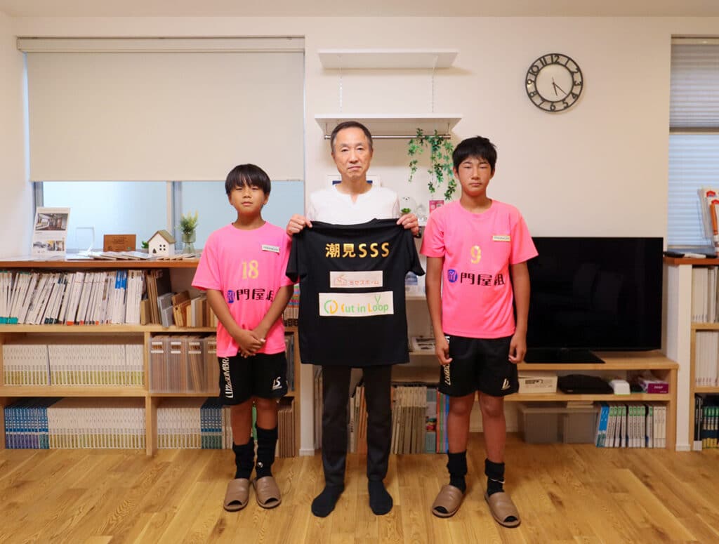 潮見サッカースポーツ少年団の選手2名とミセスホーム社長が、協賛企業ロゴ入りのチームウェアを持って記念撮影している様子